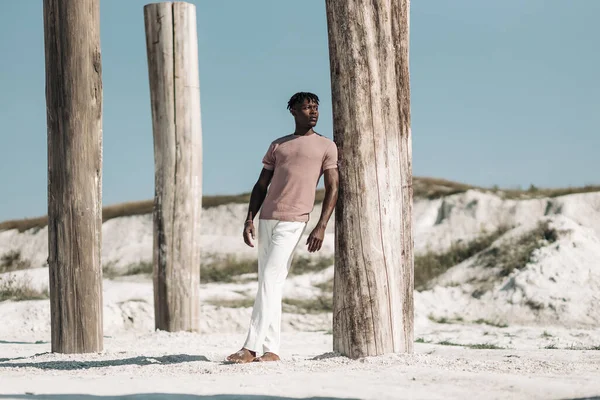 Tişört ve beyaz pantolon giyen modaya uygun siyahi bir adamın gün batımında çölde yürüyüşünün tam boy portresi.