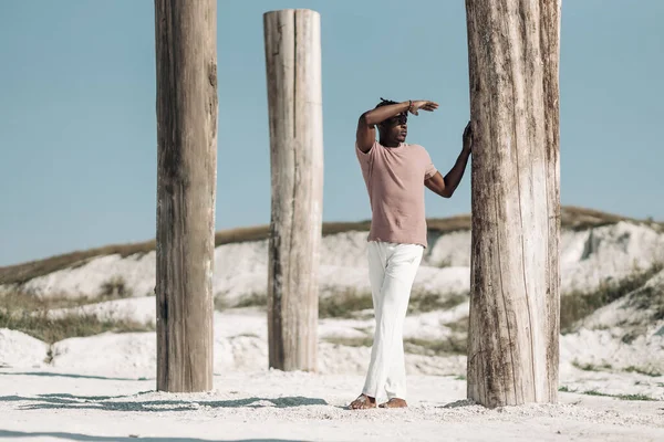 Tişört ve beyaz pantolon giyen modaya uygun siyahi bir adamın gün batımında çölde yürüyüşünün tam boy portresi.