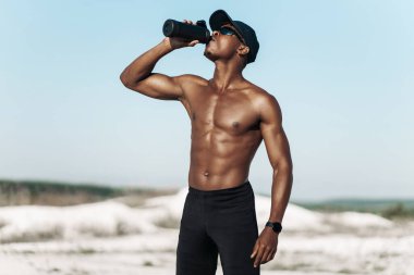 Susamış Afrikalı Amerikalı adam su şişesiyle koştuktan sonra ferahlıyor. Kaslı çıplak gövde sporcusu. Doğada dağlarda antrenman yaptıktan sonra içiyor.