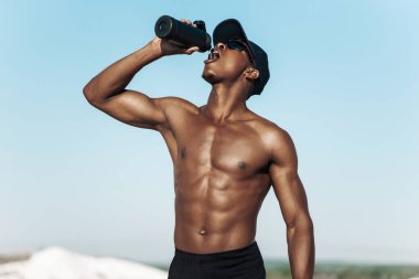 Kas yığını Afrikalı Amerikalı, içme suyu şişesi olan sporcu, dışarıda antrenmandan sonra dinlenen çekici sporcu, doğa, spor ve sağlıklı yaşam tarzı konsepti.
