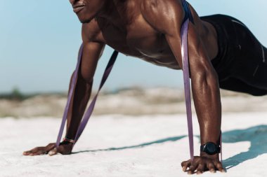 Atletik Afrikalı bir adam, elastik bantla şınav çekiyor ek zorluk olarak, açık havada, atlet lastik elastik bantları çekerek dinamik egzersizler yapıyor.