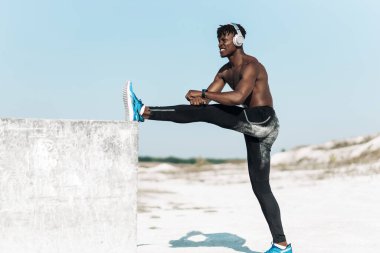 Kaslı Afrikalı Amerikalı adam kulaklıkla müzik dinliyor ve esneme egzersizleri yapıyor, dışarıda ısınıyor.