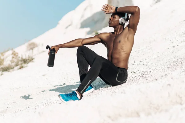 Exhausted sporty African American man, muscular man in sportswear, wearing headphones, with a bottle of drinking water, exercising outdoors in the mountains, athlete taking a break after training