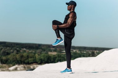 Sportif Afro-Amerikan erkek, spor giyimli, sabahın erken saatlerinde esneme hareketleri yapıyor, arka planda inanılmaz dağ manzarası var.