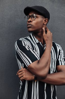 Afro-Amerikalı adam elini yüzünün önünde katladı, moda pozu verdi, düşünceli, hızlı adımlı, siyah bir duvarın arkasına yaslandı.