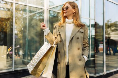 Güneş gözlüklü ve alışveriş çantalı güzel sarışın bej yağmurluk giymiş camdan bir binanın önünde duruyor. Konsept: sonbahar alışverişi, Kara Cuma, Kasım