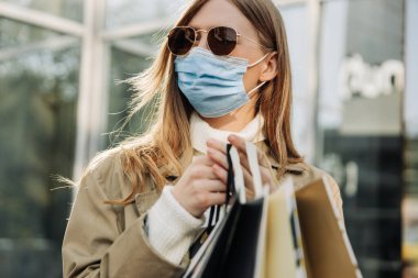 Koronavirüse karşı koruyucu maskeli şık bir kadın ve ellerinde bej yağmurluk ve güneş gözlüğü olan alışveriş torbaları. Cam bir binanın arka planına karşı. Sonbahar indirimi kavramı