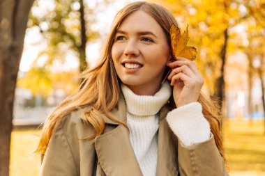 Bej renkli yağmurluk giyen güzel bir kadın ağaçtan düşmüş sarı bir yaprak gösteriyor. Sonbahar havası, ruh hali..