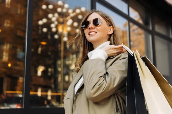 a beautiful woman in the sun of goggles in a beige raincoat and shopping bags in her hands, stands in front of a glass building, and lights. autumn theme: shopping.