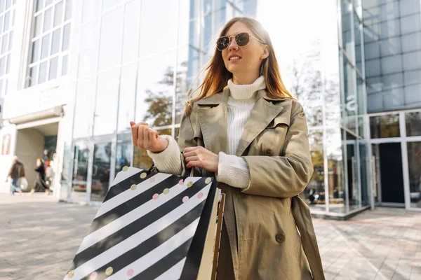 身穿米黄色雨衣戴着墨镜 手里拿着购物袋的金发欧洲女人站在一座玻璃大楼 一个购物中心 一个城市里的购物广场前 站在室外 — 图库照片