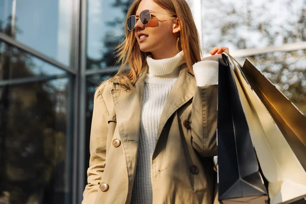 ベージュのレインコートを着た金髪のヨーロッパ人女性と彼女の手にショッピングバッグがガラスの建物の前に立っている ショッピングセンター 街のショッピング — ストック写真