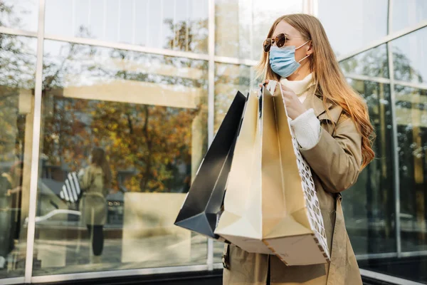 コロナウイルスから身を守るマスクをしたスタイリッシュな女性と彼女の手のショッピングバッグベージュのレインコートとサンゴーグル ガラスの建物を背景に 秋の割引の概念 — ストック写真