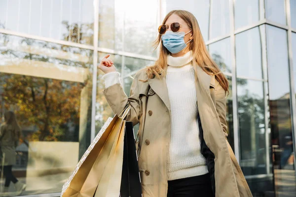 コロナウイルスから身を守るマスクをしたスタイリッシュな女性と彼女の手のショッピングバッグベージュのレインコートとサンゴーグル ガラスの建物を背景に 秋の割引の概念 — ストック写真