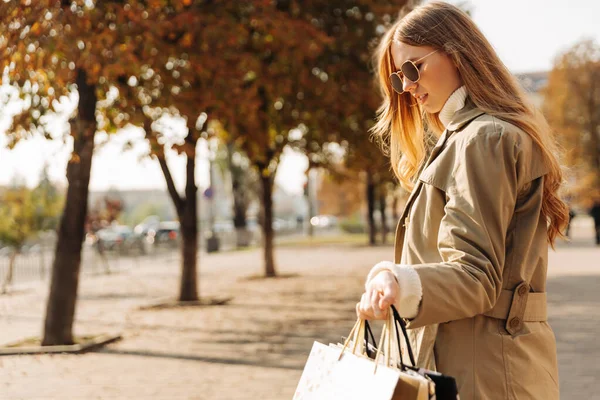 穿着米黄色雨衣的时髦女人 手里拿着购物袋的太阳镜 靠着秋天的树 打折的季节 在城市购物的女商人 — 图库照片