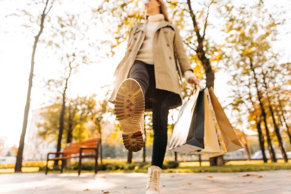 一个女人的秋鞋的特写 一个女人拿着购物袋在街上 在公园里 树都倒了 黄叶也掉了 穿着米黄色雨衣和秋靴的女人 — 图库照片