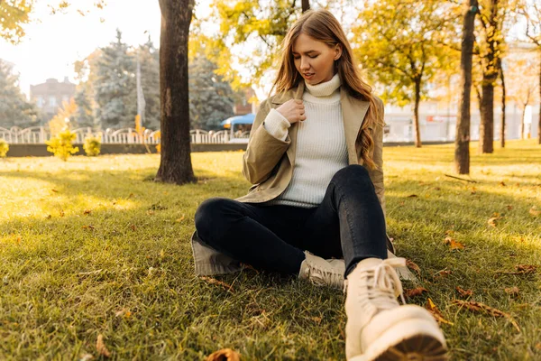 穿着米黄色雨衣的漂亮女人坐在公园的草地上 背负着秋天落叶的背景 秋天的心情 时髦的女人 — 图库照片