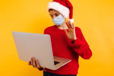 Koruyucu tıbbi maskeli, elinde dizüstü bilgisayar ve sarı arka planda Noel Baba şapkası olan bir çocuk. Odaklanarak iletişim kurar, öğretmenle çevrimiçi olur. Uzaktan öğrenme. Noel konsepti
