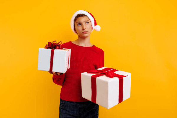 Pensive boy in a Christmas hat and a gift in his hands. Thought thinks what is inside the gifts. On a yellow background. Christmas concept