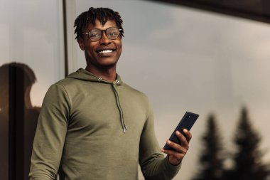 Gözlüklü Afrikalı bir adam mobil uygulama kullanıyor. Ofisin dışında mesaj atıyor. Arka planda gökdelen binaları var.