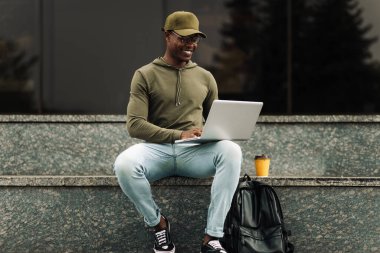 Gülümseyen Afrikalı adam, dizüstü bilgisayar kullanarak şehir merdivenlerinde oturuyor, notlar alıyor, Webinar izliyor, çevrimiçi derse giriyor, online çalışan pozitif öğrenciyi motive ediyor, laptop ekranına bakıyor, açık havada