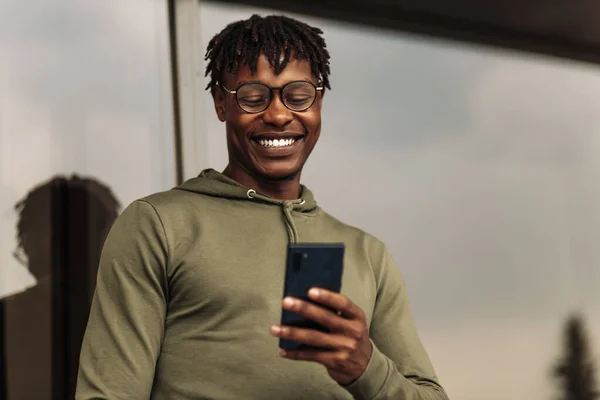 African American man with glasses using mobile app, text message outside the office in the city with skyscrapers buildings in the background, Young African man holding smartphone for business work