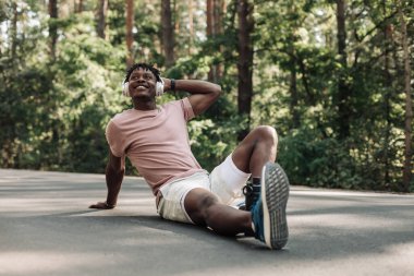 Atletik kaslı Afrikalı adam parkta koştuktan sonra kulaklıkla müzik dinliyor ormanda dışarıda antrenmanların keyfini çıkarıyor.