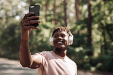 Kablosuz kulaklıklı sportif kıvırcık Afrikalı adam, parkta yaptığı çalışmadan sonra cep telefonuyla selfie çekiyor.