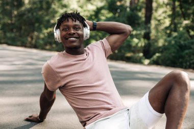 Atletik kaslı Afrikalı adam parkta koştuktan sonra kulaklıkla müzik dinliyor ormanda dışarıda antrenmanların keyfini çıkarıyor.