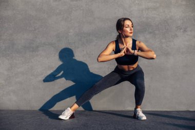 Spor giyim sektöründe spor yapan kadın spor yapıyor, esneme egzersizleri yapıyor, sokakta yoga egzersizleri yapıyor, açık hava sporları yapıyor, arka planda gri bir duvar var, şehir tarzı.