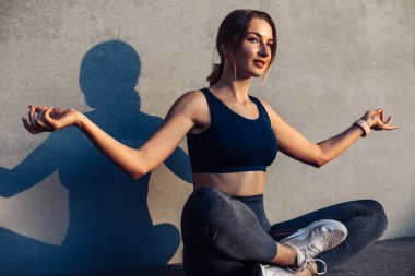 Güzel genç bir kadın yoga yapıyor, lotus pozisyonunda oturuyor, dinleniyor, şehir arka planında meditasyon yapan bir kadın, güzel bir kadın sokağın ortasında beton duvara karşı yoga yapıyor..