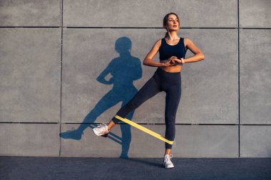 Güzel sporcu kadın direnç bandıyla egzersiz yapıyor, fitness kadını gri arka planda sarı fitness lastiğiyle bacak egzersizi yapıyor.