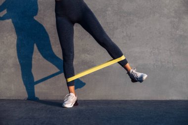 Güzel sporcu kadın direnç bandıyla egzersiz yapıyor, fitness kadını gri arka planda sarı fitness lastiğiyle bacak egzersizi yapıyor.