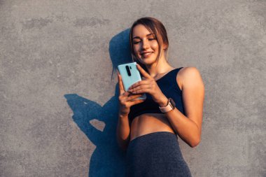 Gülümseyen kaslı formalı kadın spor giyimli, akıllı saati cep telefonu kullanan, gri şehir arka planındaki akıllı telefona bakıyor.