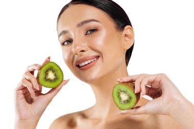 Çekici genç bir kadının portresi, parlak tenli, kivi yarımlarını gösteren, esmer, beyaz arka planda yeşil kivi yarımları tutan, güzellik ve cilt bakımı kavramı olan.