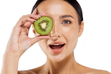 Şaşırmış genç bir kadın gözlerinin önünde kivi tutuyor, beyaz arka planda kivi dilimi olan güzel bir kadın, tropik meyveler, sağlıklı yiyecekler, güzellik, kozmetik...