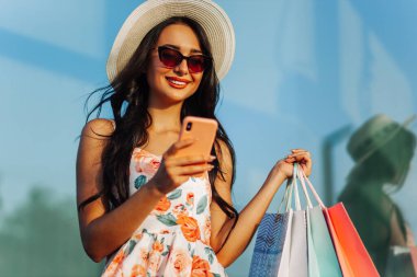 Akıllı telefon kullanarak alışveriş torbaları taşıyan güzel, genç ve şık bir kadın Kara Cuma günü indirimli alışverişler yapıyor, alışveriş merkezi yakınında, tüketim konsepti, alışveriş, Kara Cuma.