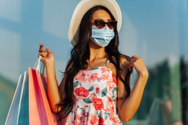 Yüzünde koruyucu tıbbi maske takan, alışveriş merkezinin yakınında alışveriş torbalarıyla gezen genç ve şık bir kadın Kara Cuma, Alışveriş, Kara Cuma, indirim, indirim, Covid-2019
