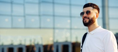 Güneşli bir günde güneş gözlüklü, beyaz gömlekli, sakallı yakışıklı bir adam gülümsüyor. Dışarıda cam binanın yanında bir adam var, Moda adamı, sokak arka planında.