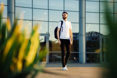 Sırt çantası ve dizüstü bilgisayarla şehirde dolaşan bir öğrenci. Güneş gözlüklü çekici genç adam güneşli bir günde sokakta yürüyor, eğitim kavramı.