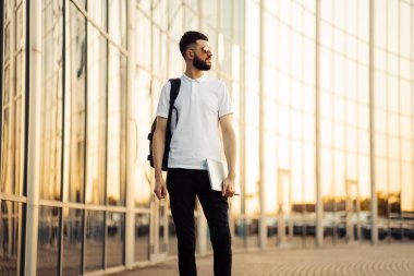 İş adamı güneş gözlüğü takıyor ve elinde dizüstü bilgisayarla şehir caddesinde yürüyen siyah bir sırt çantasıyla dışarıda bir binanın yanında yürüyen başarılı bir adam.