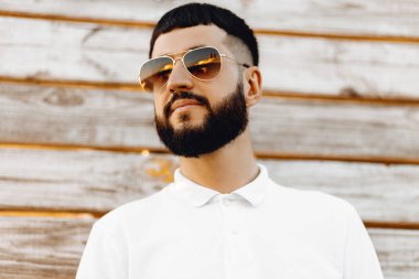 Güneş gözlüklü yakışıklı, sıradan kıyafetler giyen kameraya bakıp gülümseyen adam. Ahşap duvara yaslanmış yakışıklı bir adamın portresi.