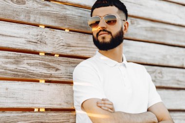 Güneş gözlüklü, sakallı, tahta bir duvarın yanında dikilen yakışıklı beyaz adam, duvarda mutlu bir şekilde gülümsüyor.