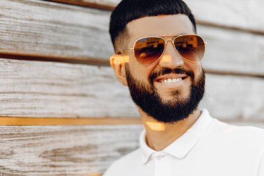 Güneş gözlüklü yakışıklı, sıradan kıyafetler giyen kameraya bakıp gülümseyen adam. Ahşap duvara yaslanmış yakışıklı bir adamın portresi.