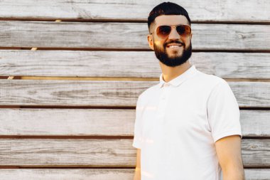 Yakışıklı, sakallı, güneş gözlüklü, tahta arka planda duruyor. Mesajınız ya da ilanınız için fotokopi odası var. Beyaz tişörtlü, dışarıda poz veren şık bir adam.