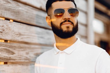 Güneş gözlüklü, sakallı, tahta bir duvarın yanında dikilen yakışıklı beyaz adam, duvarda mutlu bir şekilde gülümsüyor.