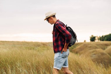 Güneş gözlüklü ve şapkalı bir gezgin gün batımında bir tarlada yürüyor. Erkek turistler yürüyüş yapar ve güzel doğanın tadını çıkarırlar. Seyahat, kamp, turizm, tatil kavramı.