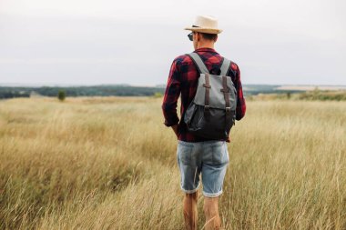 Güneş gözlüklü ve şapkalı bir gezgin gün batımında bir tarlada yürüyor. Erkek turistler yürüyüş yapar ve güzel doğanın tadını çıkarırlar. Seyahat, kamp, turizm, tatil kavramı.