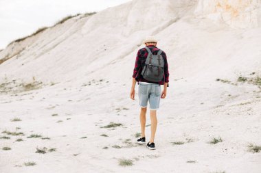 Dağların tepesinde sırt çantalı bir gezgin. Şapkalı ve güneş gözlüklü bir gezgin. Sırt çantalı bir turist bir dağın arka planında duruyor. Dağlarda ve kıyılarda seyahat ediyor.