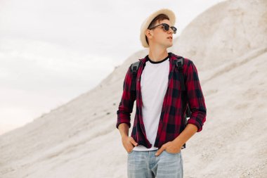 Sırt çantası ve şapkası olan güneş gözlüklü genç bir turist arka plandaki dağ manzarasına bakıyor. Doğada mutlu genç bir turistin portresi.