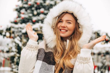 Festive Christmas fair, winter holidays concept: happy smiling woman in a scarf, catching snowflakes, posing in the city near a festive decorated Christmas tree, New Year